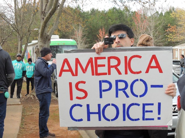A counter-protester aims a sign and a camera at a group of protesters (behind the camera). Daniel Parks, leader of extreme anti-abortion group Cities4Life, stands behind him speaking into a microphone hooked to a large speaker aimed at the clinic.