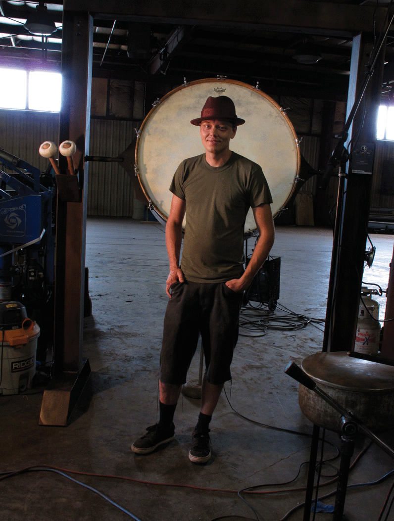 Photo by Fionn Reilly. Kris Perry stands in front of an instrument/sculpture that features a massive mounted bass drum.