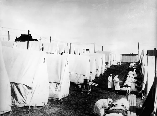 Masked doctors and nurses treat flu patients lying on cots and in outdoor tents at a hospital camp during the influenza epidemic of 1918. - CREDIT: HULTON ARCHIVE/GETTY IMAGES/TNS