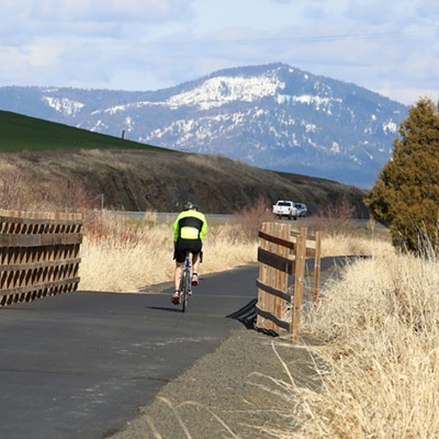 Moscow Mountain looms in the near distance as a lone ride crosses one of several bridges over Paradise Creek on the Bill Chipman Palouse Trail.  The picture was taken by Keith Collins, on Saturday, March 27, 2021, a day of floating clouds in a sunny sky over a landscape of varied greens and earth tones.
