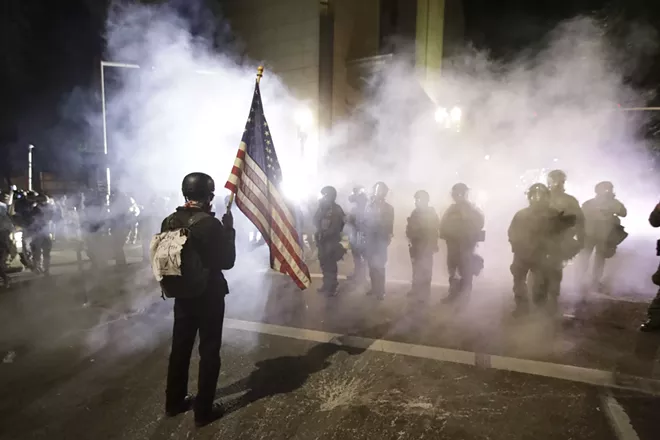 A demonstrator waves a U.S. flags in front of federal agents after tear gas is deployed during a Black Lives Matter protest at the Mark O. Hatfield United States Courthouse Thursday, July 30, 2020, in Portland, Ore. - AP PHOTO/MARCIO JOSE SANCHEZ