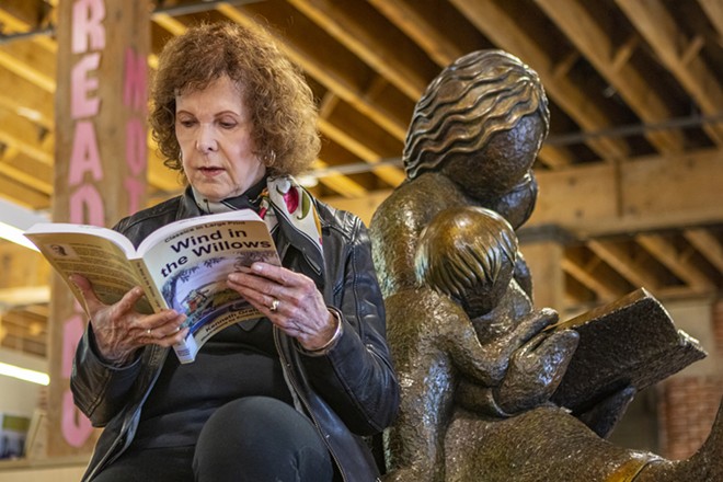 Sharon Taylor, of Lewiston, sits next to her sculpture of a mother reading to her children in the Lewiston City Library last Thursday. Taylor is working with the Lewiston City Library and the Tsceminicum Club on the - Reading Mother Challenge, in which participants receive raffle tickets to win prize baskets for reading each book imprinted along the hem of Taylor’s sculpture. - AUSTIN JOHNSON/INLAND 360
