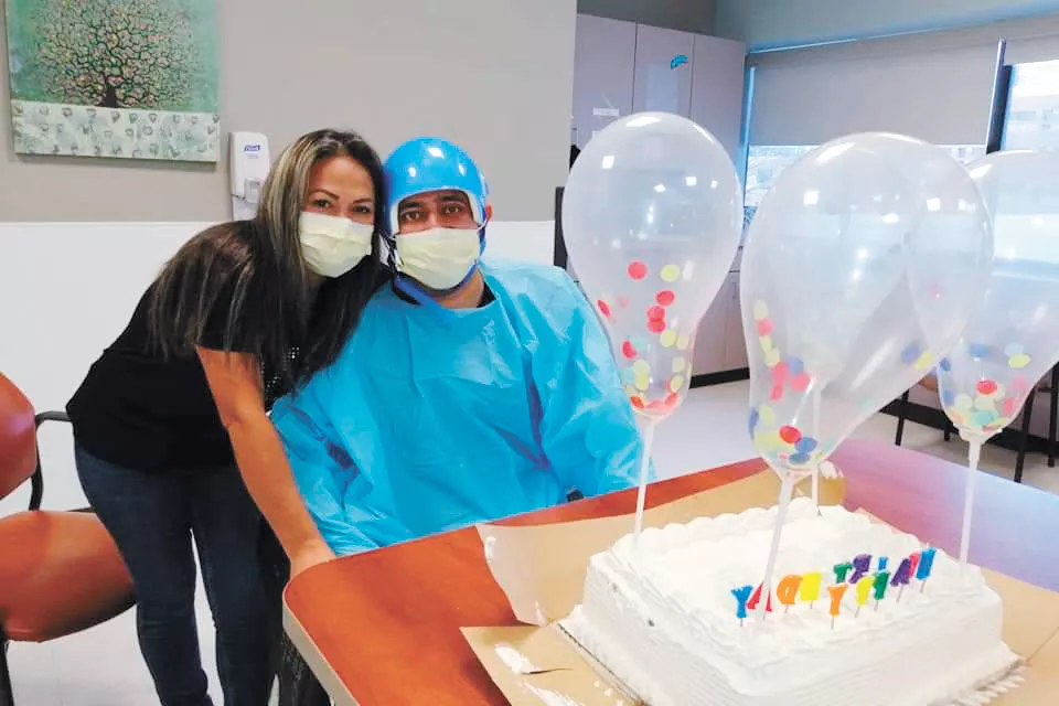 A woman stands next to a seated man wearing a blue hospital gown and a mask. At the table in front of them is a white birthday cake with clear balloons. 