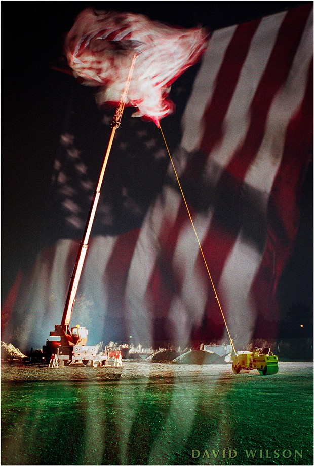 A giant U.S. flag flies beside U.S. Highway 101 between Arcata and McKinleyville, Humboldt County, California. 1991. Shot on 35mm film, this is an in-camera double exposure on a single negative; I made one exposure of the whole scene with the crane carrying the flag twisting in the wind. Then, without advancing the film, I took a telephoto shot of the flag filling the frame with its stripes flowing softly upward. The two images overlapped on the negative to produce this image. - DAVID WILSON