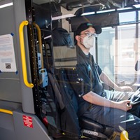 A Humboldt Transit Authority driver sits behind a Plexiglass shield wearing a facial covering, which drivers with glasses are not required to wear while buses are in motion due to safety concerns.