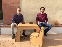 JUST ONE OF THE GUYS: Jake Disraeli (left) and Justin Farr (right), owners of The Cardboard Guys, sit with their company’s cardboard desk and chair with their logo and mascot, Cardboard Corey. - PHOTO BY REBECCA LUCAS