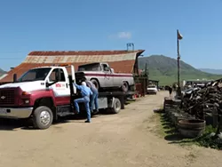 OFF THE FARM : Tow company workers endeavor to haul away one of 26 vehicles county code officials had a warrant to remove from Dan DeVaul’s Sunny Acres ranch. In the end, only six were removed. - PHOTO BY PATRICK HOWE