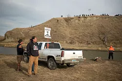 Water protectors attempting to defend Standing Rock Sioux Tribe natural resources and sacred land stand near a river as armed police line up along a hilltop near development of the Dakota Access Pipeline development. (Wade Dunstan / for Oklahoma Gazette)