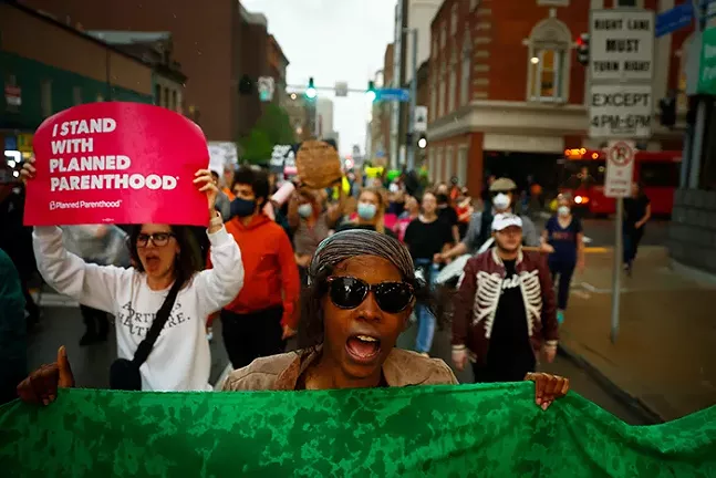 Many people march down the street shouting and holding signs. One sign reads, "I Stand With Planned Parenthood"