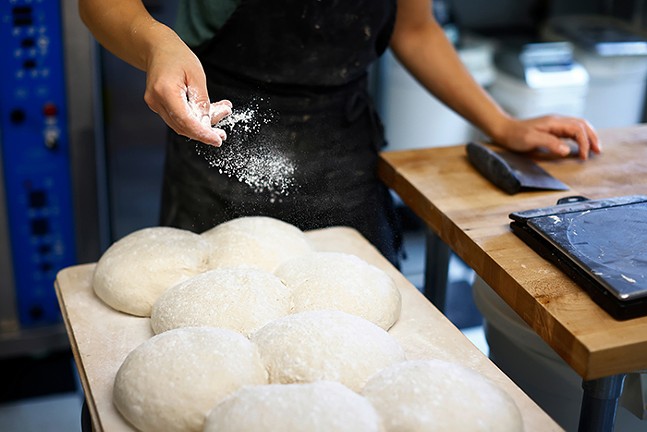A woman's hands sprinkle white flour over top of raw balls of sourdough bread sitting on a table