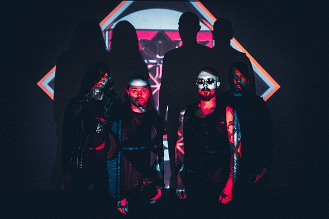 Four heavy metal musicians stand in the shadows, all partially lit by a multicolored projection.