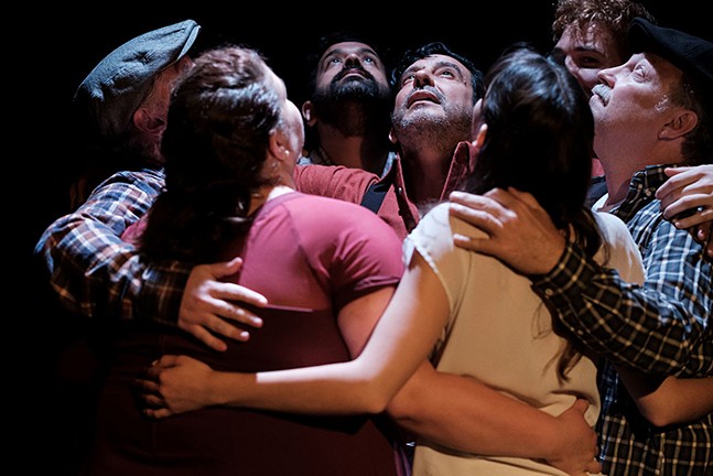 Seven people are embraced in a group hug, and all of their faces are looking upwards