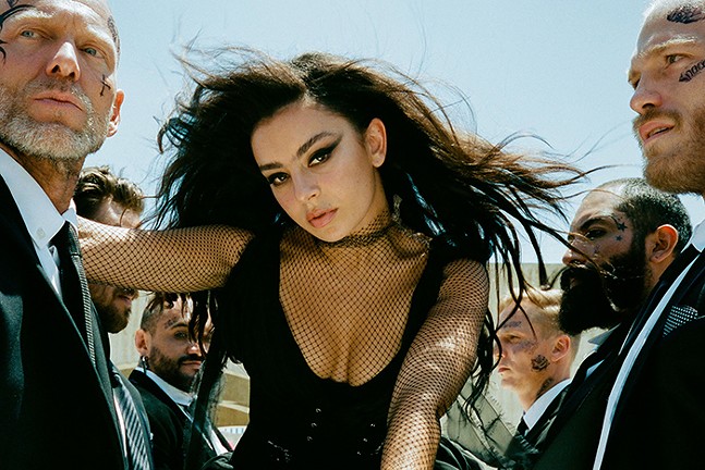 Music artist Charli XCX wears a low-cut, black mesh bodysuit and is surrounded by burly, tattooed men.