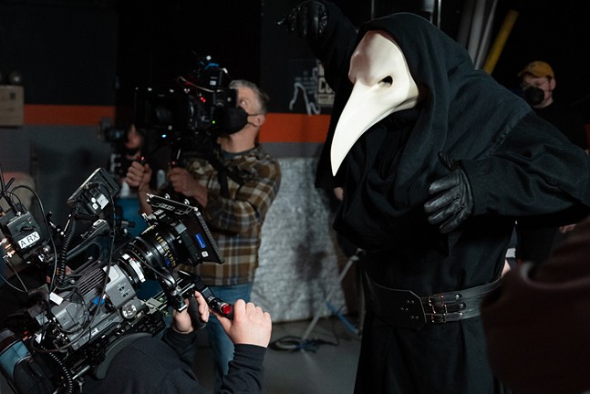 A cameraperson films up close an actor wearing robes and a giant plague mask.