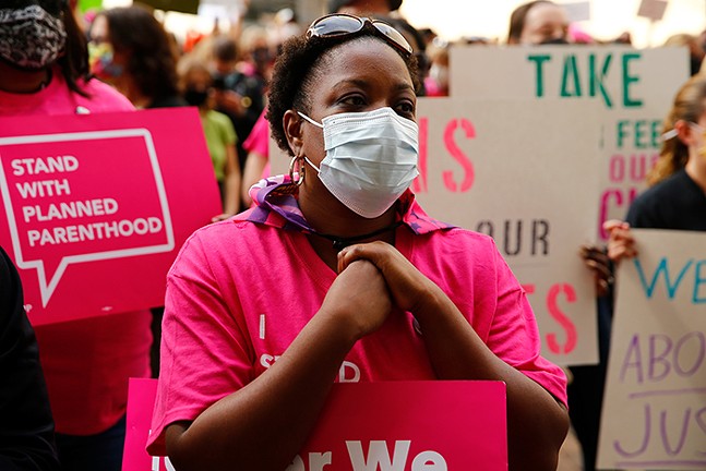 A woman wears a mask and a Planned Parenthood T-shirt in front of a crowd of people holding up signs for abortion rights