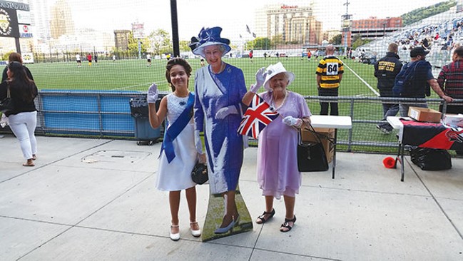 Two women (one younger, one older) dressed in fancy clothes and gloves wave the British flag next to a life-sized cardboard cut-out of Queen Elizabeth