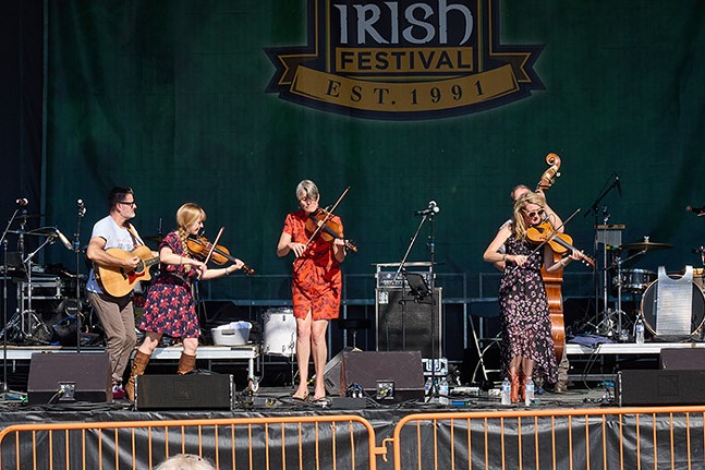 Five people on a stage playing violins, a guitar, and a bass in front of a sign that says "Irish Festival Est. 1991"