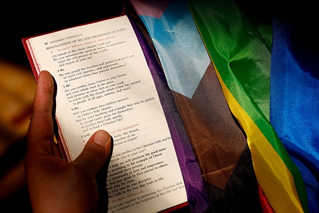 A hand holding the bible which has an LGBTQ flag draped over it