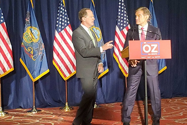 Two angry looking white men standing in front of a podium that says "Dr. OZ for Senate" and a row of American flags