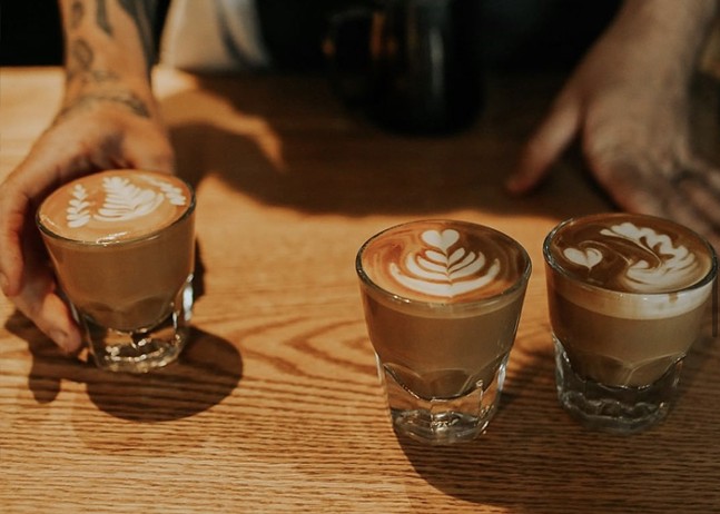 A barista serves two coffee drinks in clear glasses.