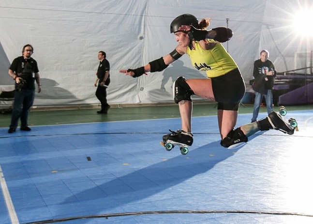 A female roller derby skater wearing a helmet and pads flies through the air as people look on.