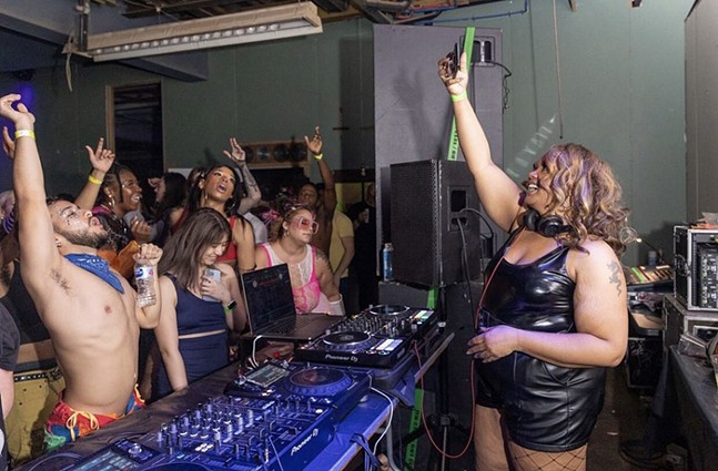 A black female DJ raises her smartphone to take a photo of the energetic dancing crowd.