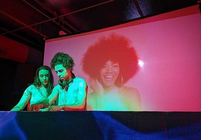 Two DJs lean over a turntable as a projection of a smiling Black woman plays behind them.