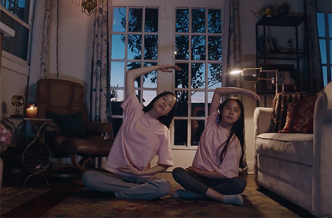 An Asian woman and girl wearing pink T-shirts sit cross-legged on the floor and do overhead stretches.