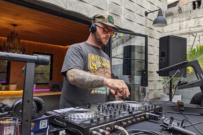 A white DJ wearing a ballcap and sunglasses turns a knob on his setup