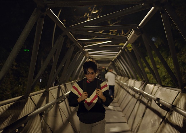 A Mexican woman wearing a striped sweatshirt and backpack looks down as she ascends a set of enclosed stairs.