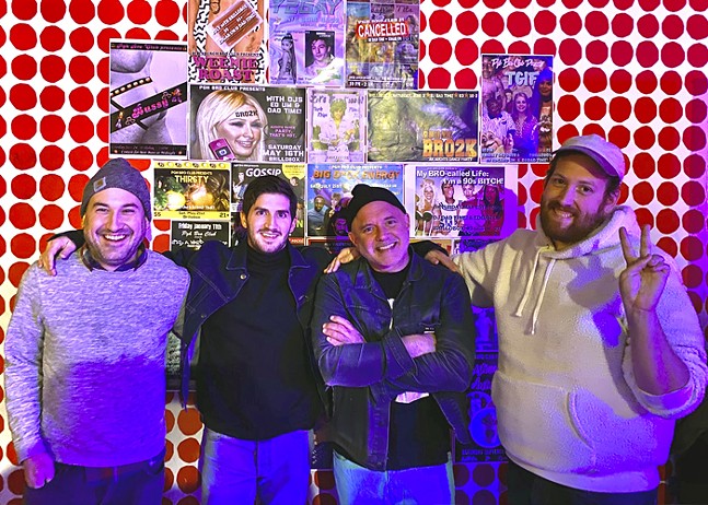 Four white DJs smile for the camera as they stand in front of a checkered red and white wall.