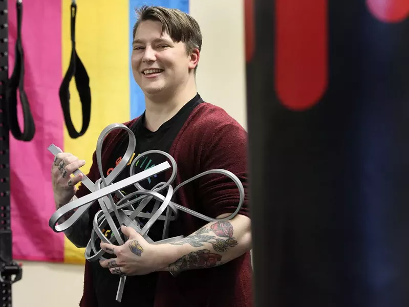 Lore McSpadden, one of the founders of Positive Force Movement, stands with a sculpture they made by bending steel with nothing but the strength and leverage of their own body. “I can’t do it again for a few more weeks, though,” McSpadden says. “I had top surgery last month.” - PHOTO BY MAX SCHULTE