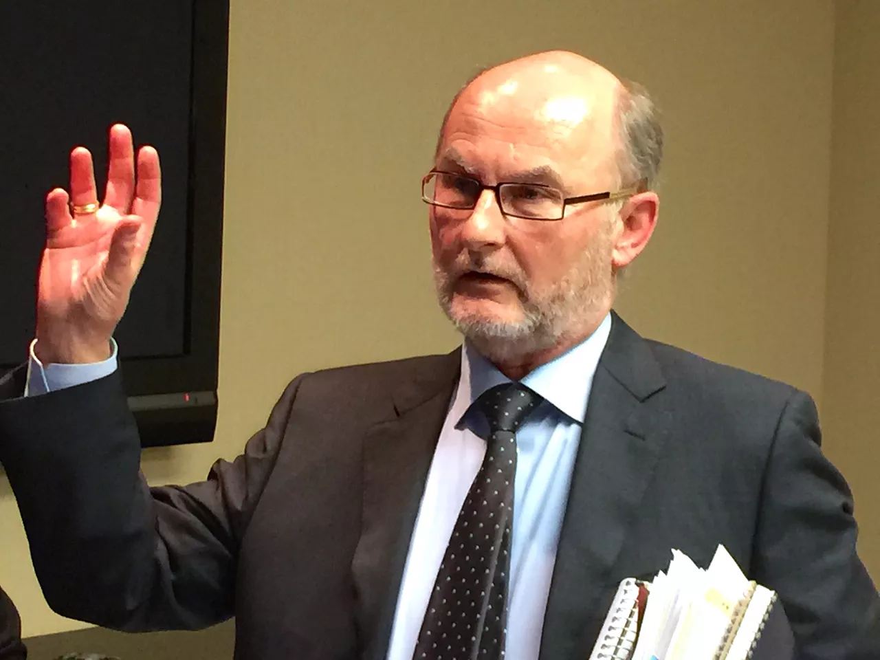 A white man with glasses holding a bunch of papers talking with someone who isn't pictured.